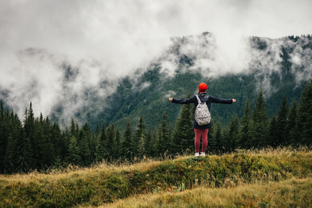 Female backpacker feeling the moment, celebrating success in wild nature of the Carpathians, shot from back, looking at fir trees and low clouds (fog) in Autumn.の写真素材