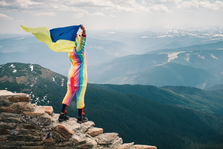 Traveling to Ukraine. Hiker in a unicorn suit standing on top of the mountain with a flag in her raised hands, looking at Carpathian landscape on a Spring day. Wanderlust  travel concept.の写真素材