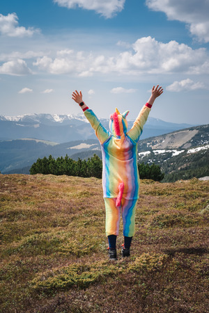 Funny travel, female hiker in a unicorn suit celebrating success, standing on top of the mountain overlooking Carpathian landscape on a Spring day. Wanderlust concept.の写真素材
