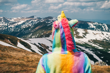 Portrait shot from back of a female hiker clad in unicorn suit looking at mountain peaks covered with snow on a sunny day in Spring. Carpathians. Wanderlust travel concept.の写真素材