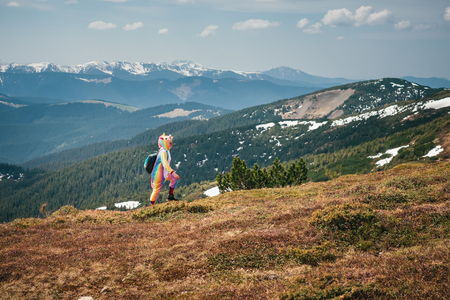 Full-length portrait of a female backpacker in a unicorn suit walking across Carpathian mountains covered with snow leftovers on a sunny Spring day. Wanderlust travel concept.の写真素材