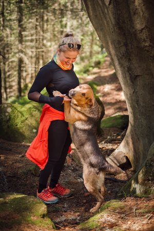 Young blonde female hiker in sporty outfit playing with a dog in a forest trip on a sunny day. Concept friendshipの写真素材