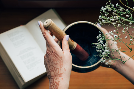 Female hands covered with henna tattoo holding tibetan singing bowl over the open book on a flower background. Meditation and inspiration moodの写真素材