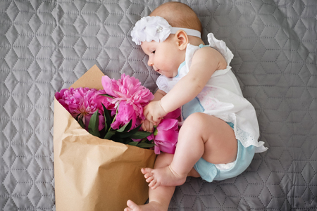 Happy newborn baby lying on the bed and playing with ping bouquet of pion flowersの写真素材