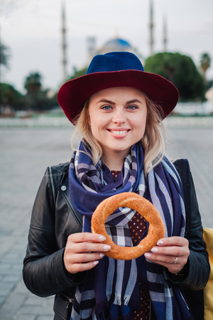 Happy beautiful tourist woman in hat and dress walking on Istanbul street and testing traditional turkish simit or bagel food, Turkeyの写真素材