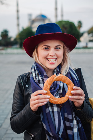 Happy beautiful tourist woman in hat and dress walking on Istanbul street and testing traditional turkish simit or bagel food, Turkeyの写真素材