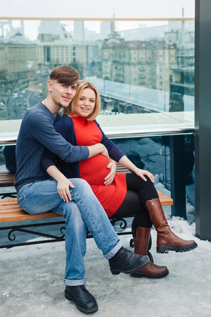 Family with pregnant woman siting on the roof top of the skyscraper in winter city back. Young wife with husband enjoy spend time togetherの写真素材