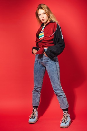 Positive retro styled fashionable woman portrait in sport jacket and jeans on red background. Fashion back to 80s - 90s new trend conceptの写真素材