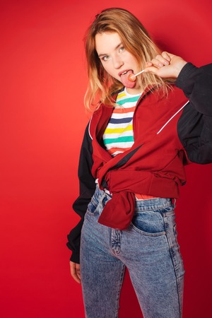 Portrait of retro styled fashionable woman in sport jacket and jeans lick lollipop on red background. Fashion back to 80s - 90s new trend conceptの写真素材