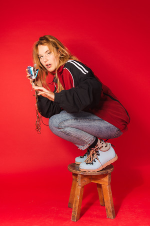 Vibrant retro styled fashionable woman in sport jacket and jeans with vintage cassette player on red background. Fashion back to 80s - 90s new trend conceptの写真素材