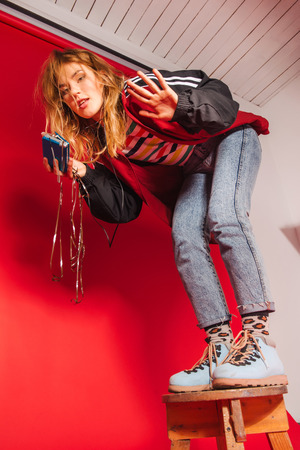 Vibrant retro styled fashionable woman in sport jacket and jeans with vintage cassette player on red background. Fashion back to 80s - 90s new trend conceptの写真素材