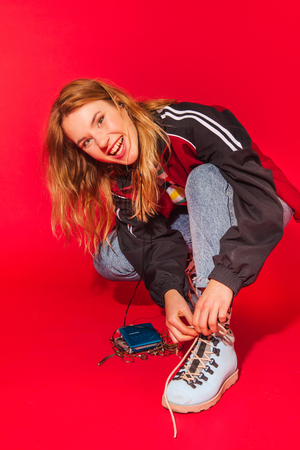Positive retro styled fashionable woman in sport jacket and jeans with vintage cassette player on red background. Fashion back to 80s - 90s new trend conceptの写真素材