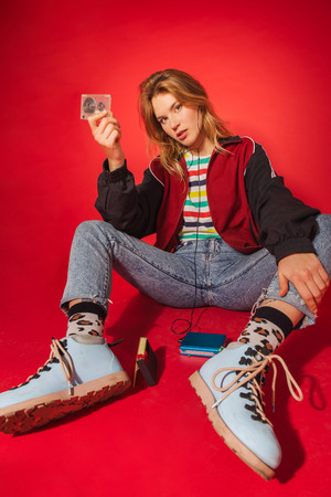 Funky retro styled fashionable woman in sport jacket and jeans with vintage cassette player on red background. Fashion back to 80s - 90s new trend conceptの写真素材