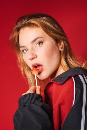 Portrait of retro styled fashionable woman in sport jacket and jeans lick lollipop on red background. Fashion back to 80s - 90s new trend conceptの写真素材