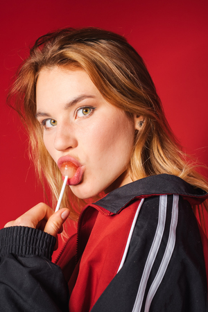 Portrait of retro styled fashionable woman in sport jacket and jeans lick lollipop on red background. Fashion back to 80s - 90s new trend conceptの写真素材