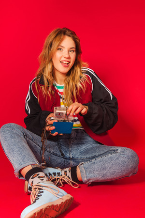Positive retro styled fashionable woman in sport jacket and jeans with vintage cassette player on red background. Fashion back to 80s - 90s new trend conceptの写真素材