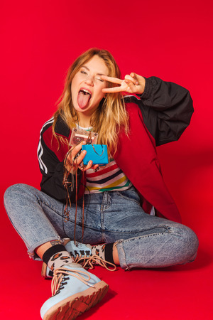 Vibrant retro styled fashionable woman in sport jacket and jeans with vintage cassette player on red background. Fashion back to 80s - 90s new trend conceptの写真素材