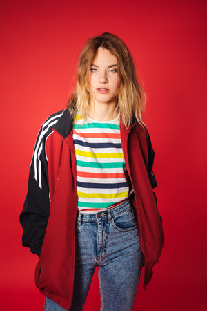 Positive retro styled fashionable woman portrait in sport jacket and jeans on red background. Fashion back to 80s - 90s new trend conceptの写真素材