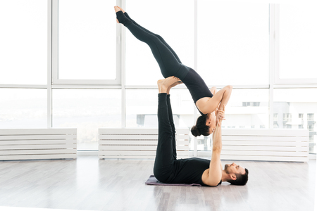 Beautiful young  woman practice acro yoga exercise with man partner  in white fitness studio with big windows on backgroundの写真素材