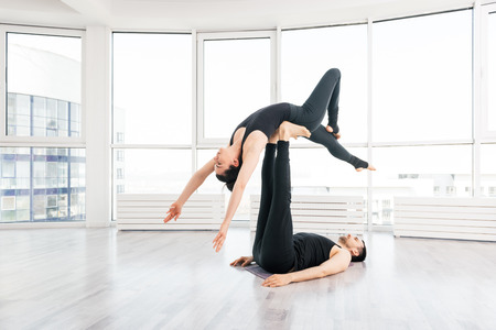 Beautiful young man and woman practice pair acro yoga exercise in white fitness studio together  with big windows on background. Man lying on wood floor and balancing womann in his feetの写真素材