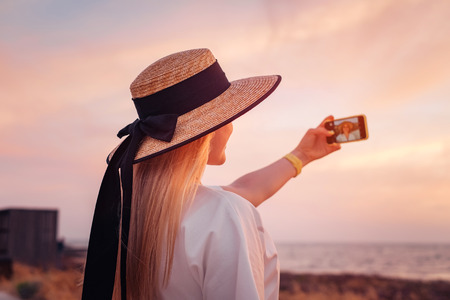 Beautiful blonde woman in white dress travel to Cyprus and enjoying summer sea sunset and take selfie on mobile phone. Freedom conceptの写真素材