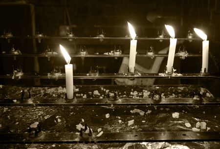 4 burning white candles in a church, the foreground candle is in focus and the others are out of focus, the flames are pointing to the leftの写真素材
