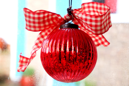 close up of shining red christmas bauble with red and white material bow on top, it is hanging in a window and the colourful back ground is out of focus behindの写真素材