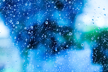 Hundreds of white rain drops on a glass window with a blurred background of blue, green, and white colours of varying shadesの写真素材