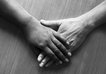 looking down on two fifty year old female hands and lower arms placed ontop of each other, the top hand is Asian and the bottom hand is caucasian, black and white photograph, the background is a wooden table top.の写真素材