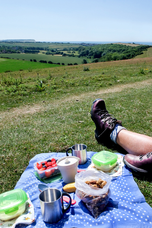 A breakfast picnic of muesli fruit yogurt and coffee laid out on a blue and white spotted table cloth on the grass on a hill looking over rolling fields and the sea in the background on a sunny day a persons leg and hiking boot are in the left corner.の写真素材