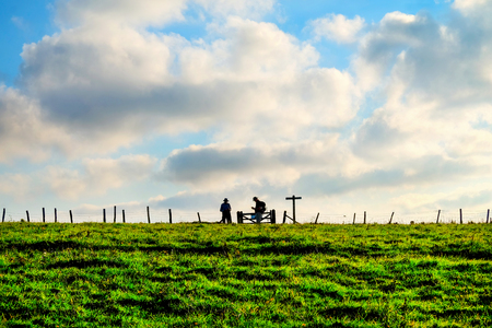 A green field in the distance are two hikers silhouetted standing at a gate next to a sign post with a wooden fence running across the centre of the image behind is a large expanse of blue sky with cloudsの写真素材