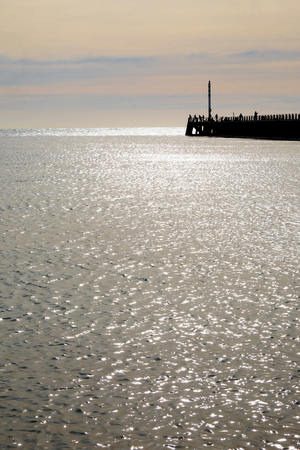Newhaven, East Sussex, UK, United Kingdom, harbour wall jutting out from the right hand side, it is silhouetted black with the silhouettes of people fishing on the end the pier in front the sea is glistening silver from the sun and the sun has set and the sky is glowing yellow and red, vertical format plenty of copy space in both the sea and skyの写真素材