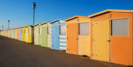 A row of twenty brightly coloured beach huts the nearest one is the largest the row has a diminishing perspective they are coloured orange, green yellow blue behind them is blue, there are two street lamps in the row the nearest one has a seagull sitting on it. Seaford, Sussex, UKの写真素材