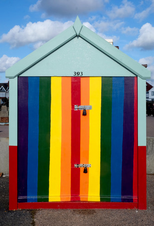 A single beach hut with a dramatic colourful striped door the stripes are blue, green yellow, oragnge, red  the sky behind is blue with fluffy white clouds the hut is sitting on a concrete promenadeの写真素材