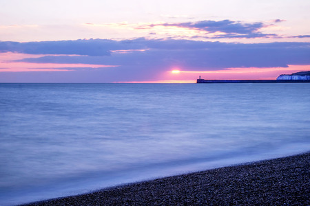 The sea is calm with movement, in the distance there is a harbour wall with a lighthouse on the end of it behind are some cliffs, the sky has scattered clouds and is glowing pink from the setting sun, the sun is setting just above the lighthouse, Newhaven, East Sussex, England, United Kingdom, UK,の写真素材