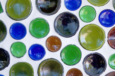 Fifty circular glass bottles have been reused as building materials and the bottom of the bottles are built into a white wall, the bottles have just the bottom of the circular glass showing, they are blue, green and brownの写真素材