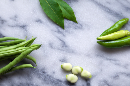 green beans, broad beans clove leaves and green chilli on a white and blue marble chopping board, the vegetables are arranged around the edges of the board leaving space in the middle of the chopping board for copy space, still life, horizontal formatの写真素材