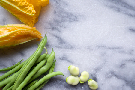 raw yellow courgette flowers, green beans and broad beans on a white and blue marble chopping board, the vegetables are arranged on the left side of the image leaving space on the right side of the chopping board for copy space, still life, horizontal formatの写真素材