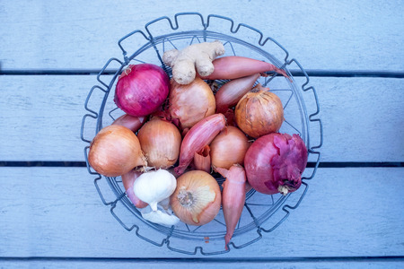 A circular wire metal bowl full of fouteen raw onions of different varieties, raw garlic and ginger, it is on a grey painted wooden table top, still life style.の写真素材