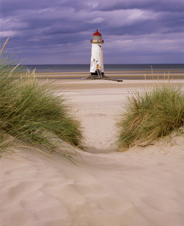 An old white light house with a bright red roof sitting on a sandy beach with a stone pathway infront, the sea is in the distance and a big blue sky with white swirling clouds is behind the light house, Talacre lighthouse, Prestatyn, North Wales, United Kingdom, UK,の写真素材