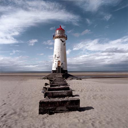 An old white light house with a bright red roof sitting on a sandy beach with a stone pathway infront, the sea is in the distance and a big blue sky with white swirling clouds is behind the light house, Talacre lighthouse, tの写真素材