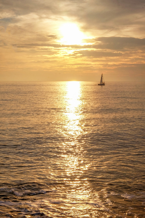 A sailing boat with three people on it silhouetted in the distance in a calm orange glowing seamless sea and sky at dusk, the sun is low in the sky and shining through the clouds casting a long highlight on the sea, vertical format,の写真素材