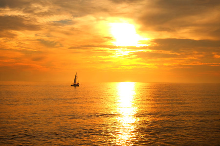 A sailing boat with three people on it silhouetted in the distance in a calm orange glowing seamless sea the sun is shining through the clouds and cast a long hilghlight on the seaの写真素材