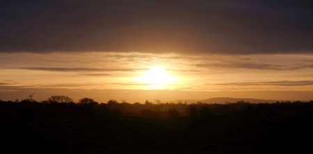 The sun is rising over the Sussex countryside just infront of the low sun are lines of trees and hills back lit by the light, the sky above the sun has a band of dark cloud, Long horizontal format, the image has a band of dark in the forground a band of yellow in the middle and a band of dark at the top, ideal for copy spaceの写真素材