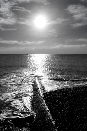 Black and white photograph of the low winter sun shining onto a flat calm sea, a long high light reflecting on the sea and across a long sea break half on the beach half in the sea casting highlights and shadows and silhouettes add an artist look to the imageの写真素材