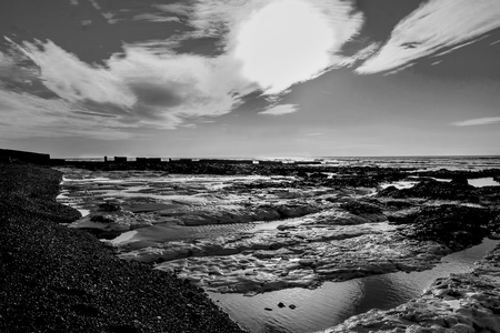 A black and white photograph of rock pools next to a pebble beach behind is the sea and a clear sky with white clouds, the sun is low in the sky back lighting the rock pools with highlights and shadows, the image is artistic, Ovingdean, East Sussex, United Kingdomの写真素材