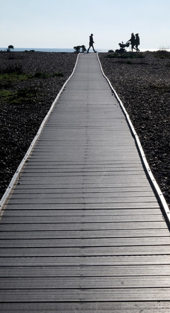 Three silhouetted unrecognisable people and a baby in a pram walking across the end of a long straight wooden pathway with a pebble beach on both sides a small line of sea and blue sky is behindの写真素材