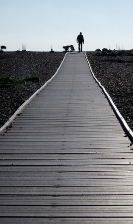 Silhouette of a man walking towards the end of a long straight wooden pathway with a diminishing perspective on a pebble beach on both sides at the end is a clear blue skyの写真素材