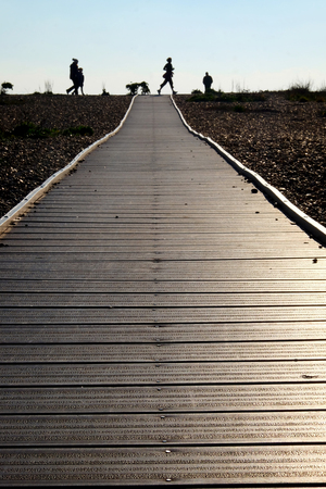 Looking down a long straight wooden pathway with a pebble beach on both sides at the end are unrecognizable people walking and jogging silhouetted by the clear blue sky behindの写真素材