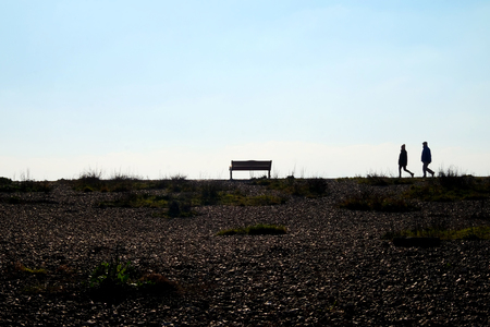 In the foreground is a pebble beach at the end of the beach are two unrecognizable people walking together as a couple they are siluoetted by the sun and sky behind, they are walking towards a park bench which is also siluoetted, Shoreham, East Sussex, UK,の写真素材